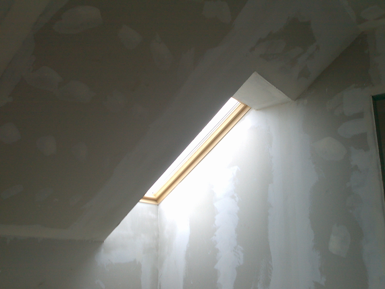 Wnętrze poddasza z oknem dachowym w trakcie prac wykończeniowych, ściany i sufit pokryte gładzią gipsową, widoczne łączenia płyt gipsowo-kartonowych.